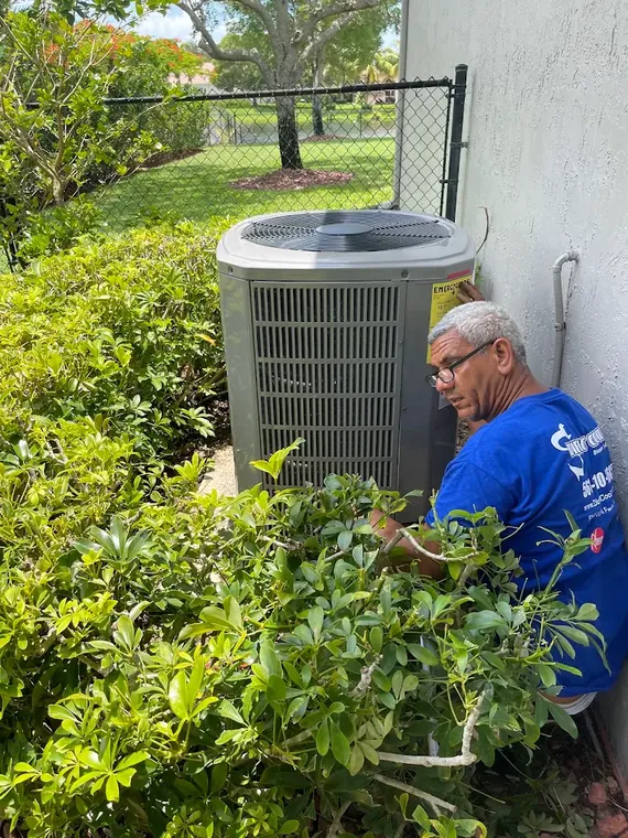 A Direct Cooling technician works on the service panel of a residential AC condenser set between shrubs and the house wall. Direct Cooling technician servicing an outdoor AC condenser behind landscaping